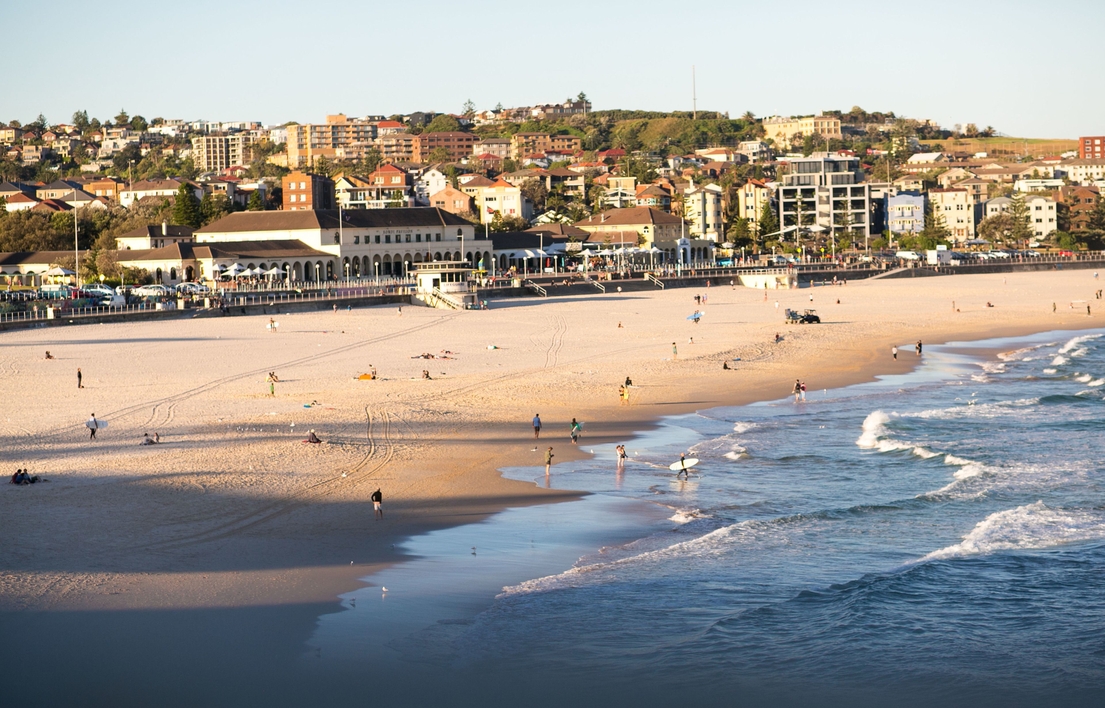 A beach with people walking and surfing, a pier with buildings, and a hillside with houses.