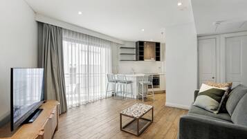 A modern living room with a flat-screen TV, a grey sofa, and a coffee table. There is a dining area with a white table and chairs, and a kitchen with a white countertop and cabinets.