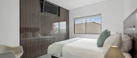 A modern bedroom with a large bed, a bedside table with a lamp, a chair, a wardrobe, and a window with a view of a house.