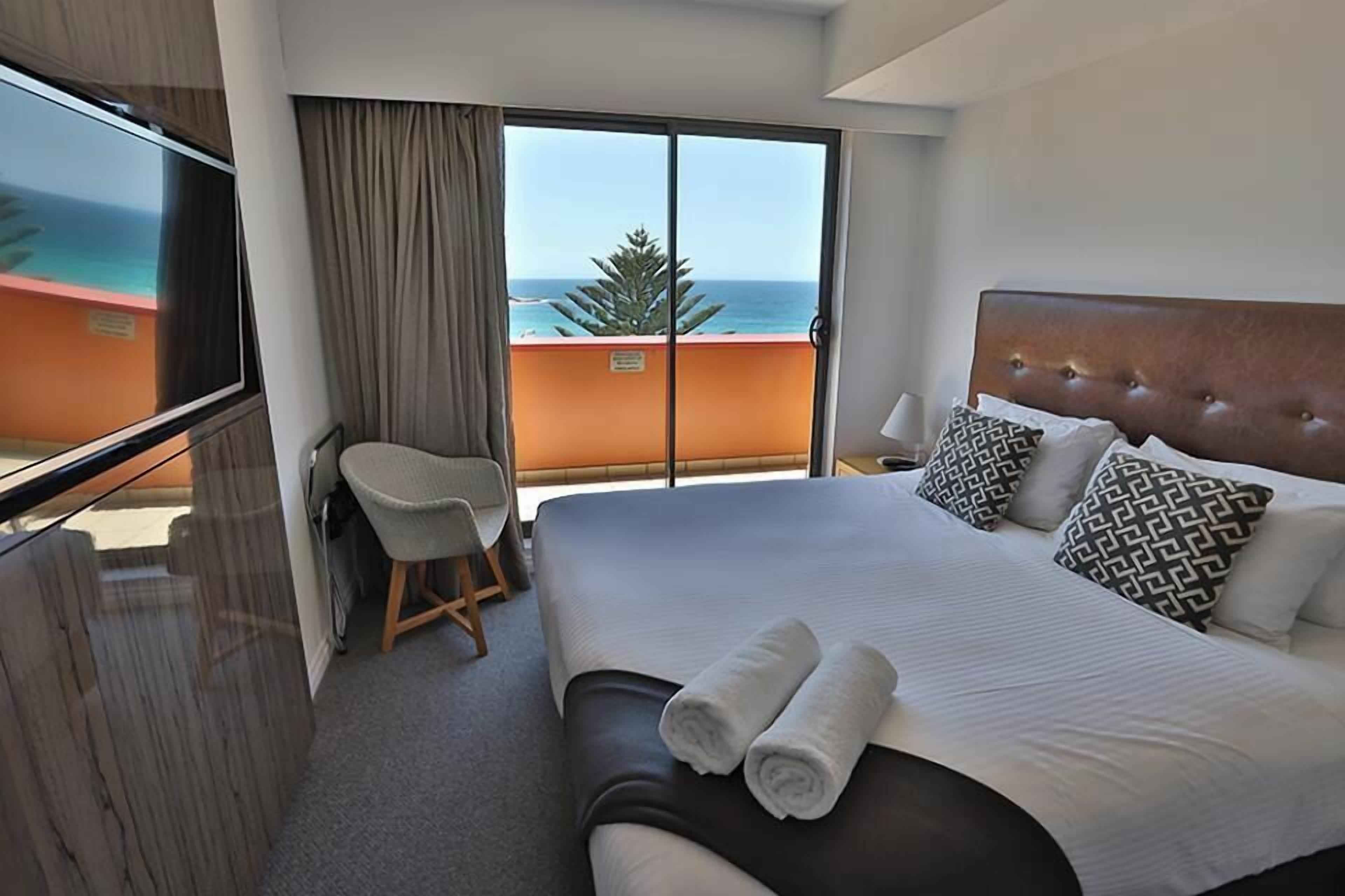 A hotel room with a large bed, a chair, a TV, and a balcony with a view of the ocean.