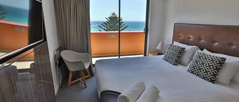 A hotel room with a large bed, a chair, a TV, and a balcony with a view of the ocean.
