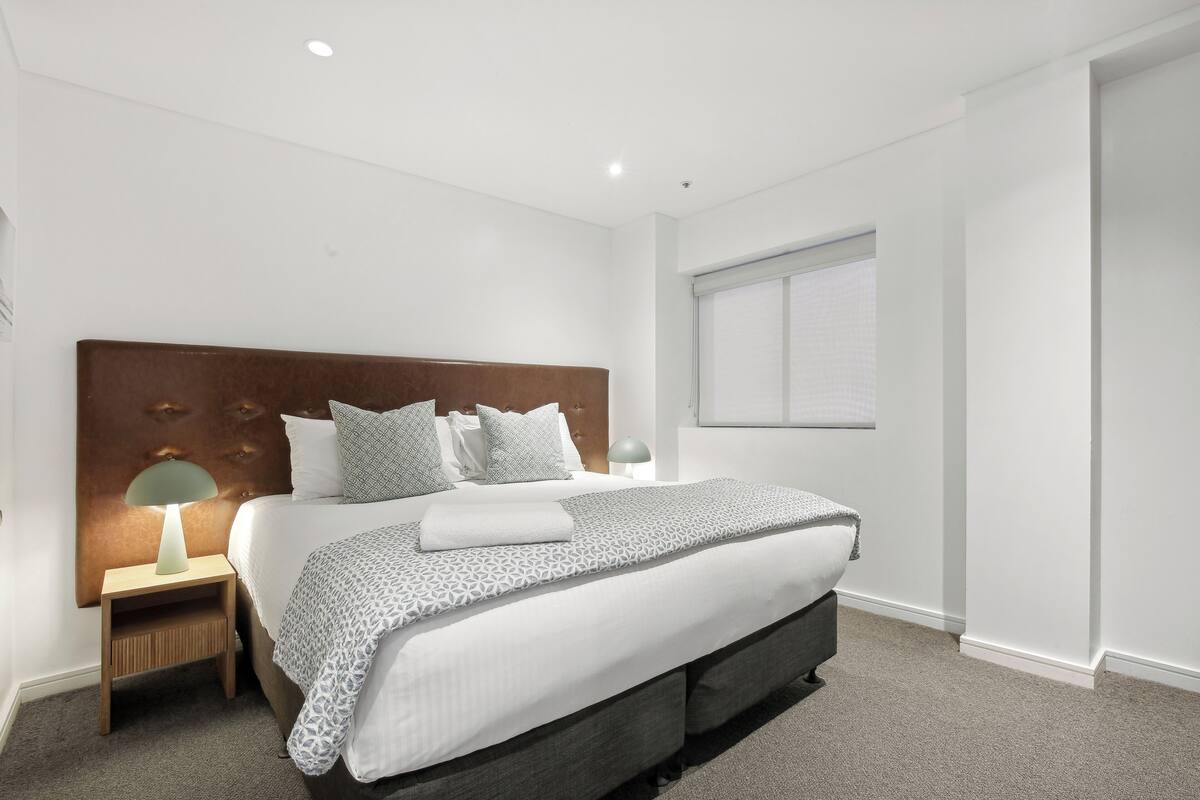 A modern hotel room with a large bed, a wooden headboard, a bedside table with a lamp, and a window with blinds.