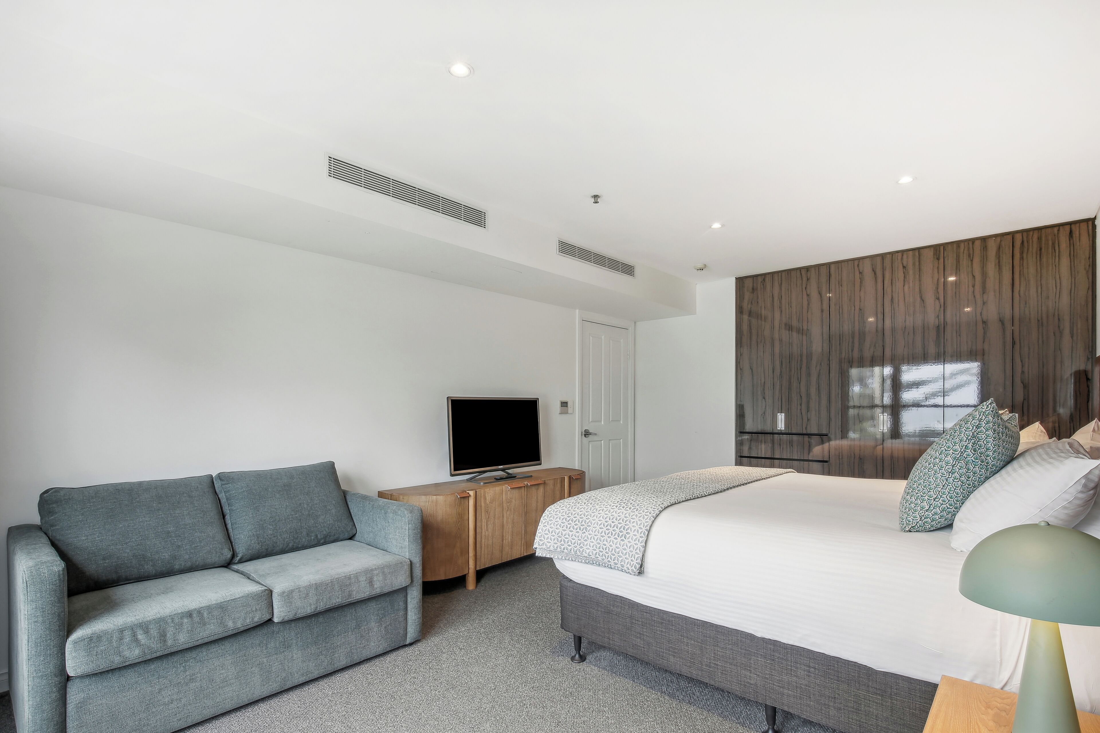 A modern hotel room with a large bed, a grey sofa, a TV on a wooden cabinet, and a window with curtains.