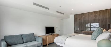 A modern hotel room with a large bed, a grey sofa, a TV on a wooden cabinet, and a window with curtains.
