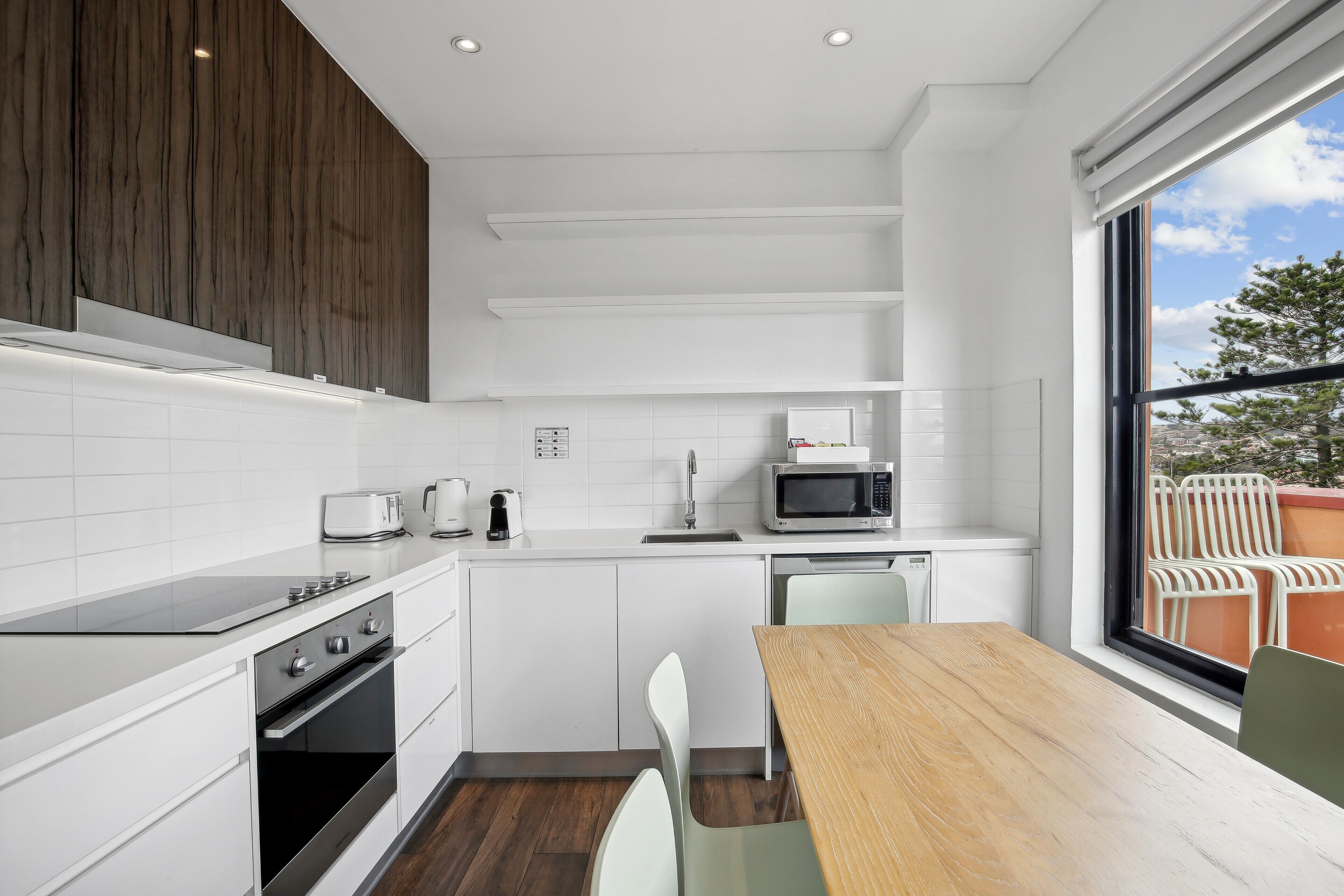 Una cocina moderna con armarios blancos, una mesa de comedor de madera y vistas a una zona de estar exterior a través de un amplio ventanal.
