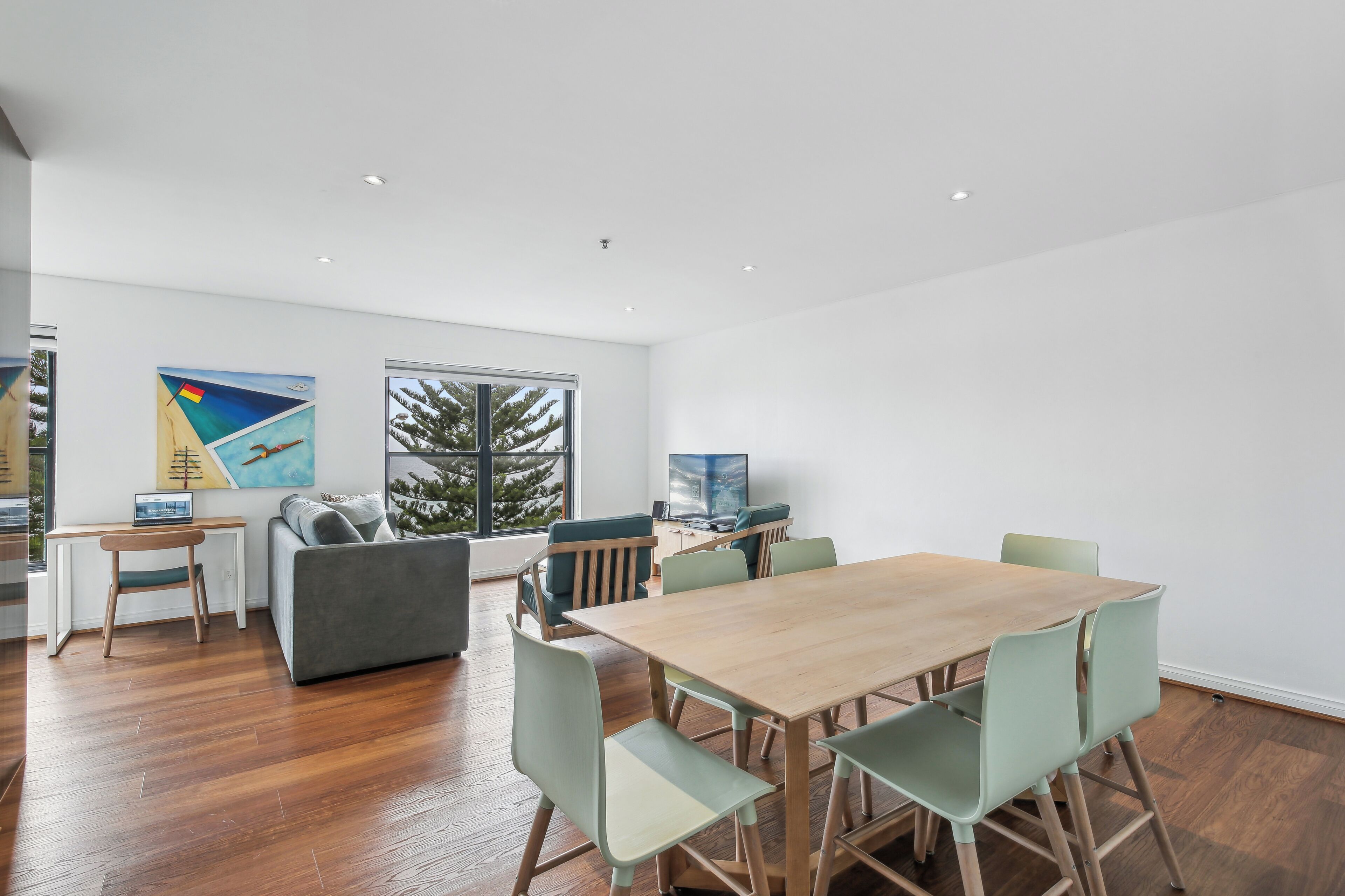 A modern living room with a wooden dining table, green chairs, a grey sofa, and a TV.