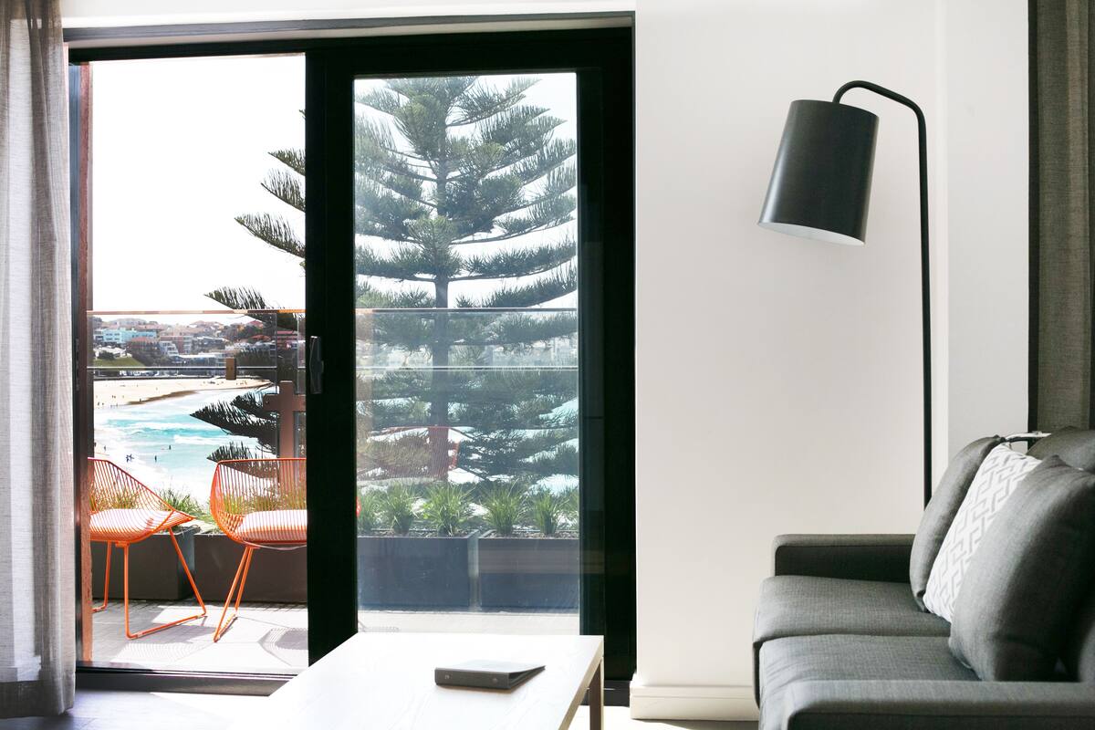 A modern living room with a grey sofa, an orange chair, a table, and a large window offering a view of a pool and trees.