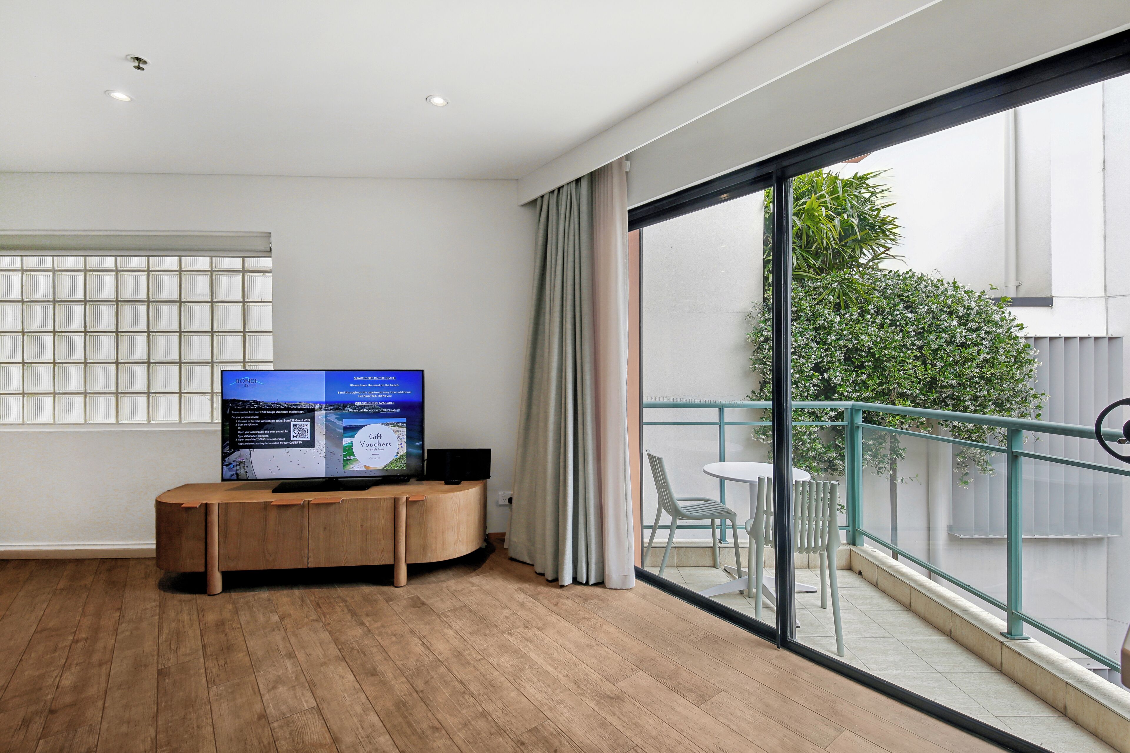 A modern room with a wooden desk, a flat-screen TV, a large sliding glass door leading to a balcony with a view of greenery.
