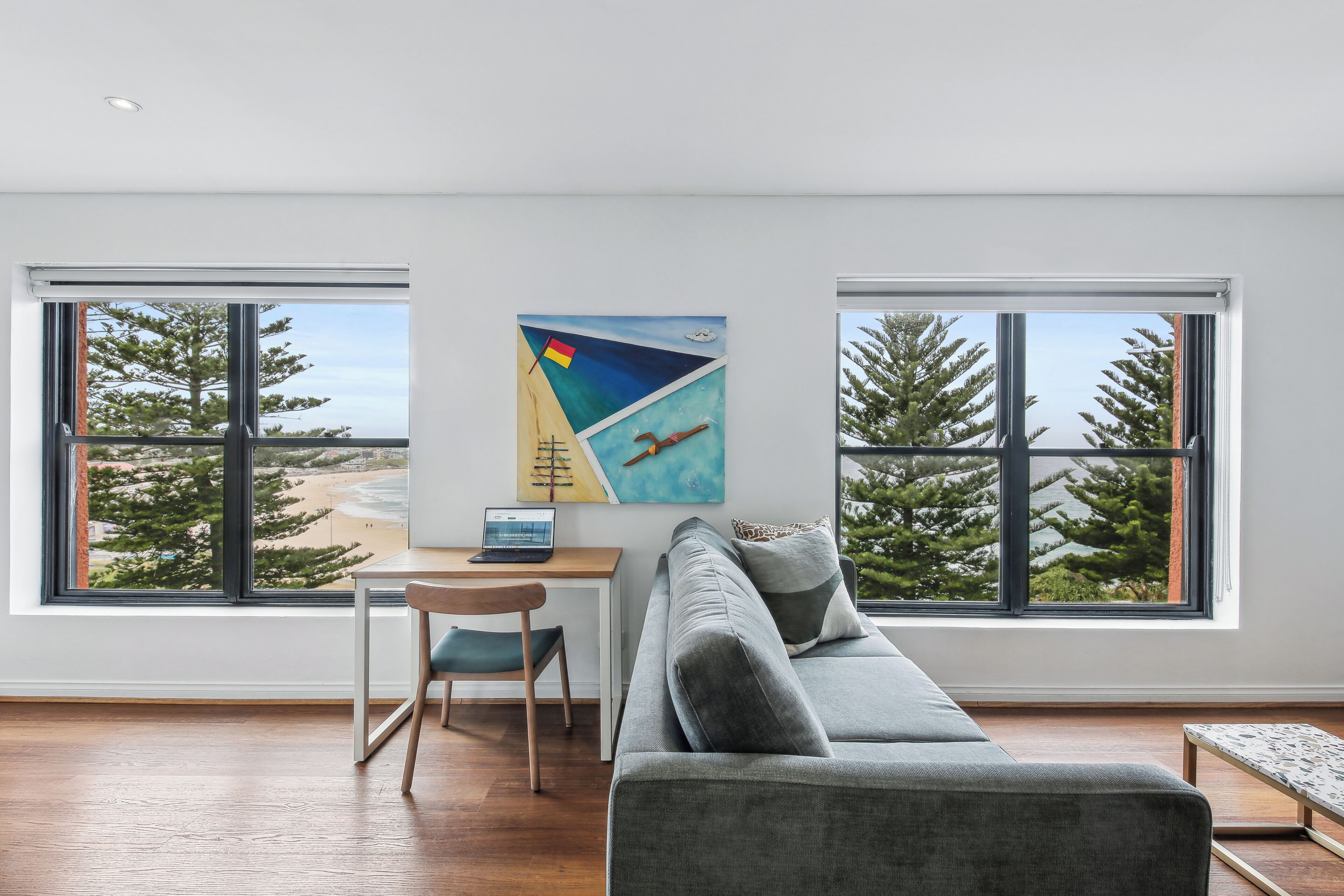A modern living room with a grey sofa, a wooden desk, and a laptop. There are two large windows with a view of trees and a beach.