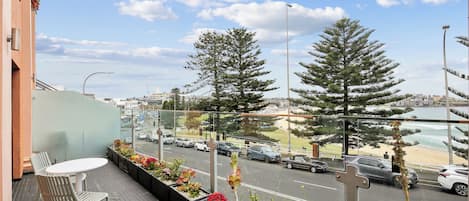 Un balcón con una mesa y sillas, con vista a una calle con coches y una playa a lo lejos.