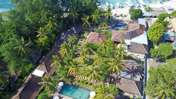 Vista aérea de un resort con piscina rodeada de palmeras, una playa con barcos y varias edificaciones con techos de tejas.