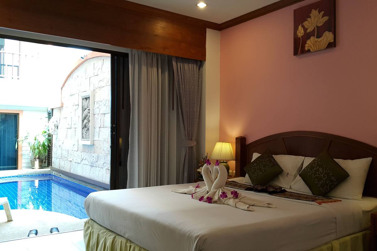 A hotel room with a bed, two bedside lamps, a nightstand with a vase of flowers, a view of a swimming pool, and a painting on the wall.
