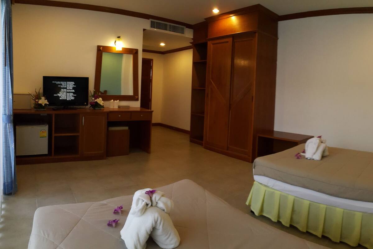 A hotel room with a bed, a desk with a computer, a wardrobe, and a bedside table with a lamp and flowers.
