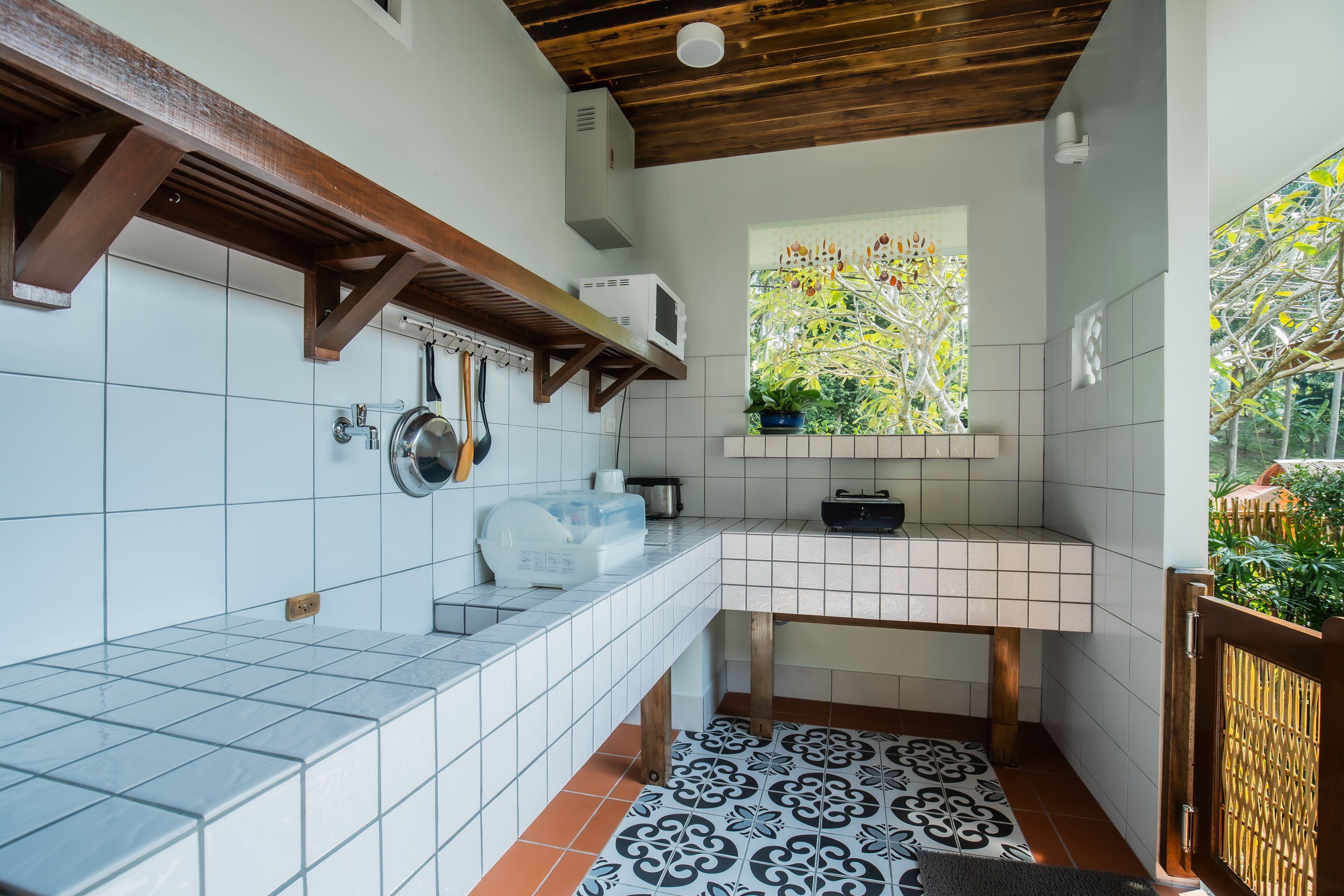 A kitchen with white tiles, a wooden ceiling, a window with a view of greenery, and a tiled floor with a black and white pattern.