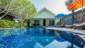 A swimming pool with lounge chairs and umbrellas, surrounded by greenery and a building with a tiled facade. - Kata Tranquil Villa (Karon)