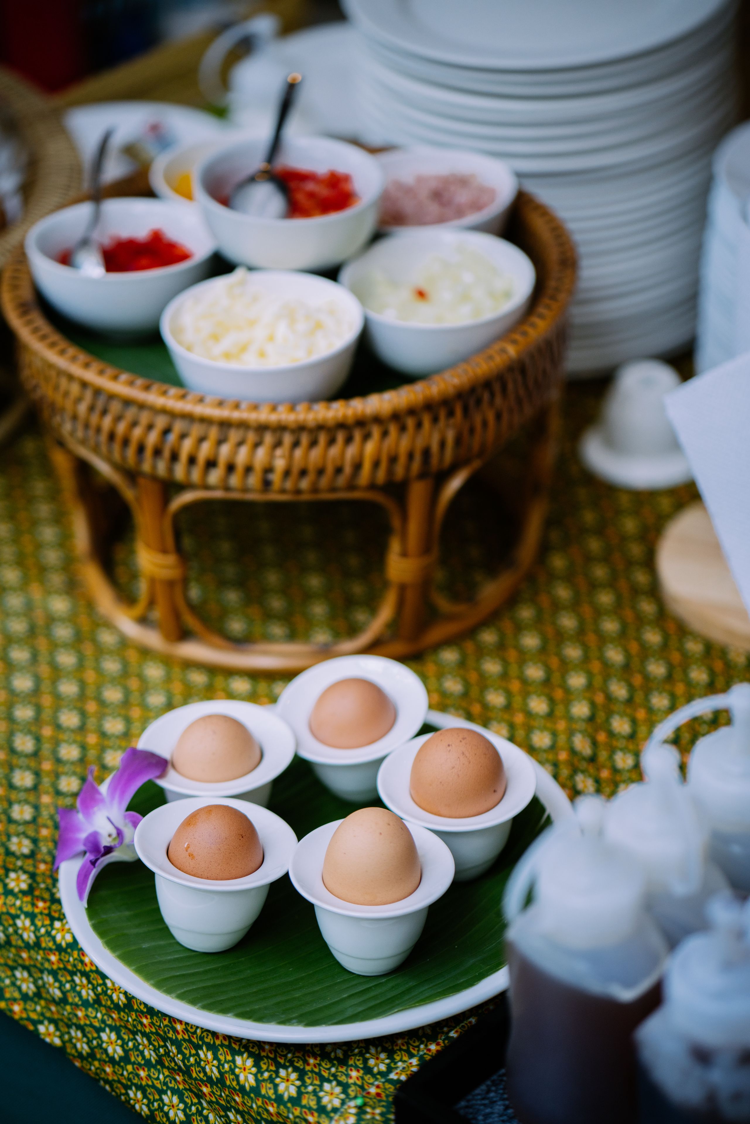 Una mesa con una cesta de mimbre que contiene recipientes de condimentos, un plato con huevos y una orquídea púrpura decorativa.