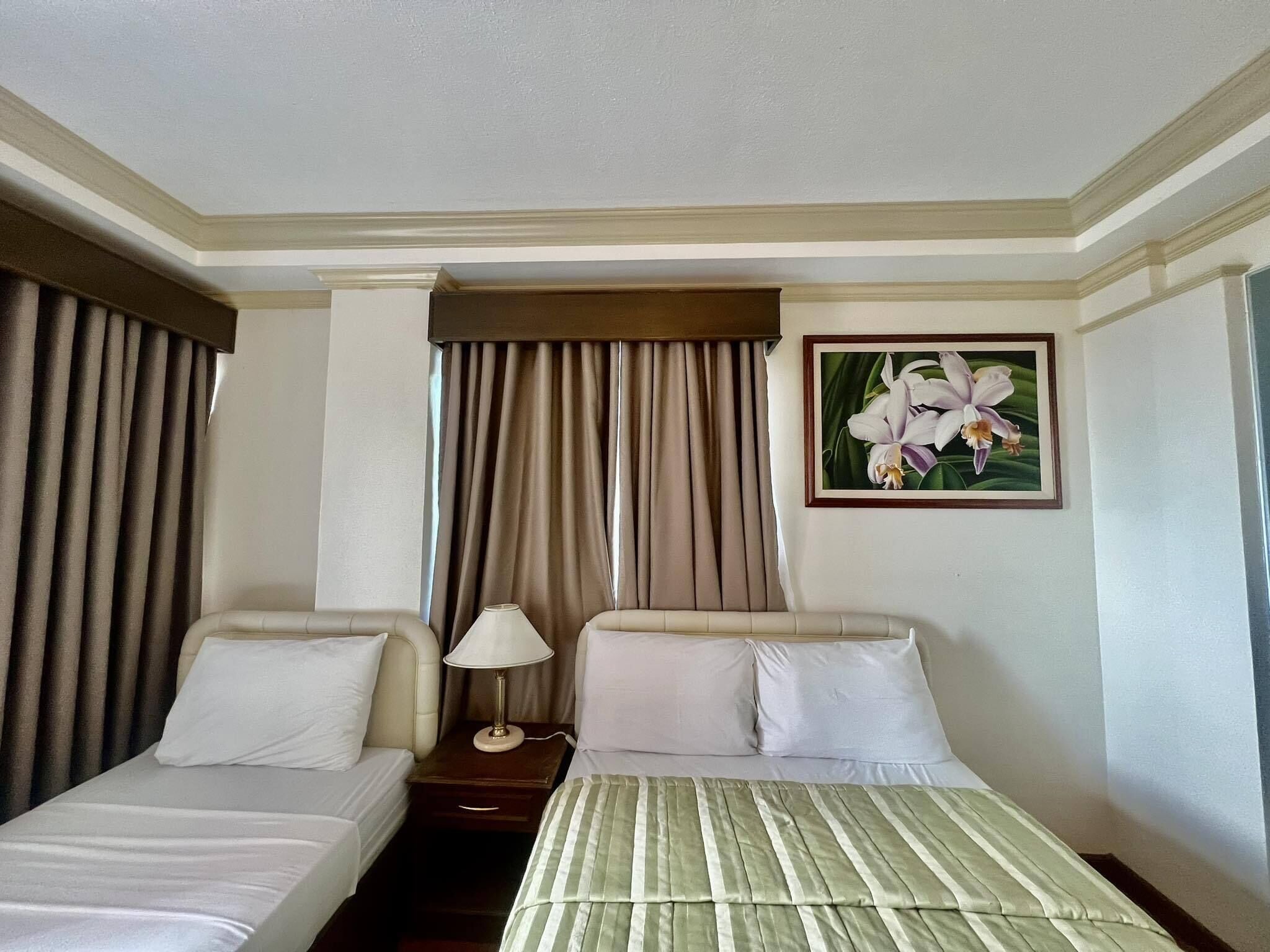 A hotel room with two single beds, a nightstand with a lamp, a painting of flowers on the wall, and a window with curtains.