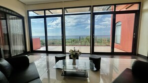 A spacious room with a large sliding glass door leading to a balcony with a view of the ocean and greenery.