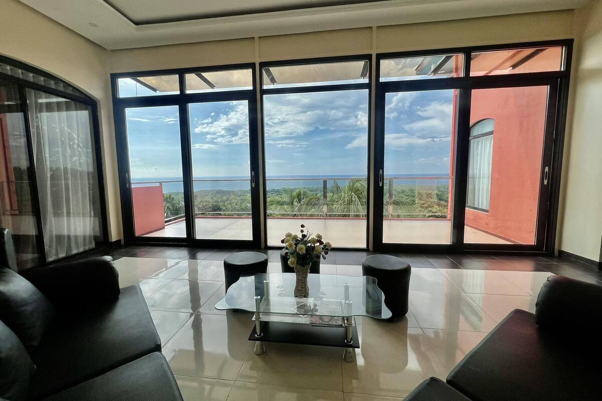 A spacious room with a large sliding glass door leading to a balcony with a view of the ocean and greenery.