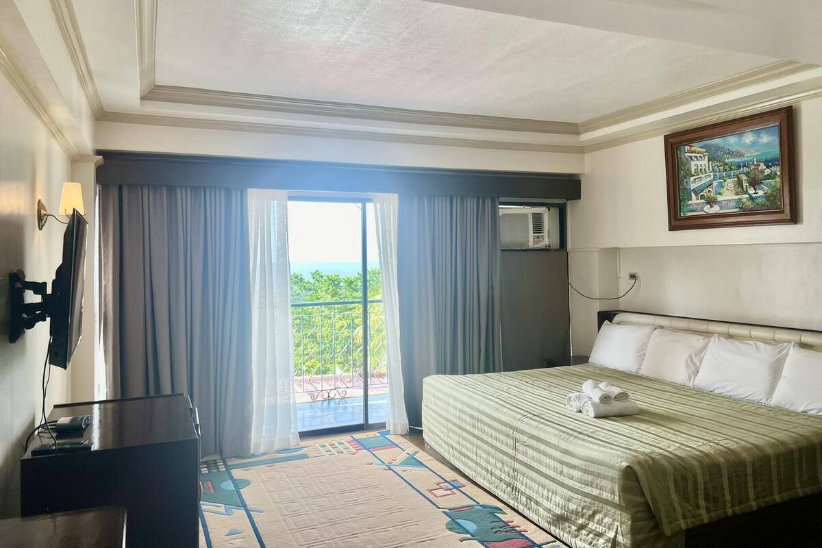 A hotel room with a bed, a desk, a TV, and a balcony with a view of the ocean.