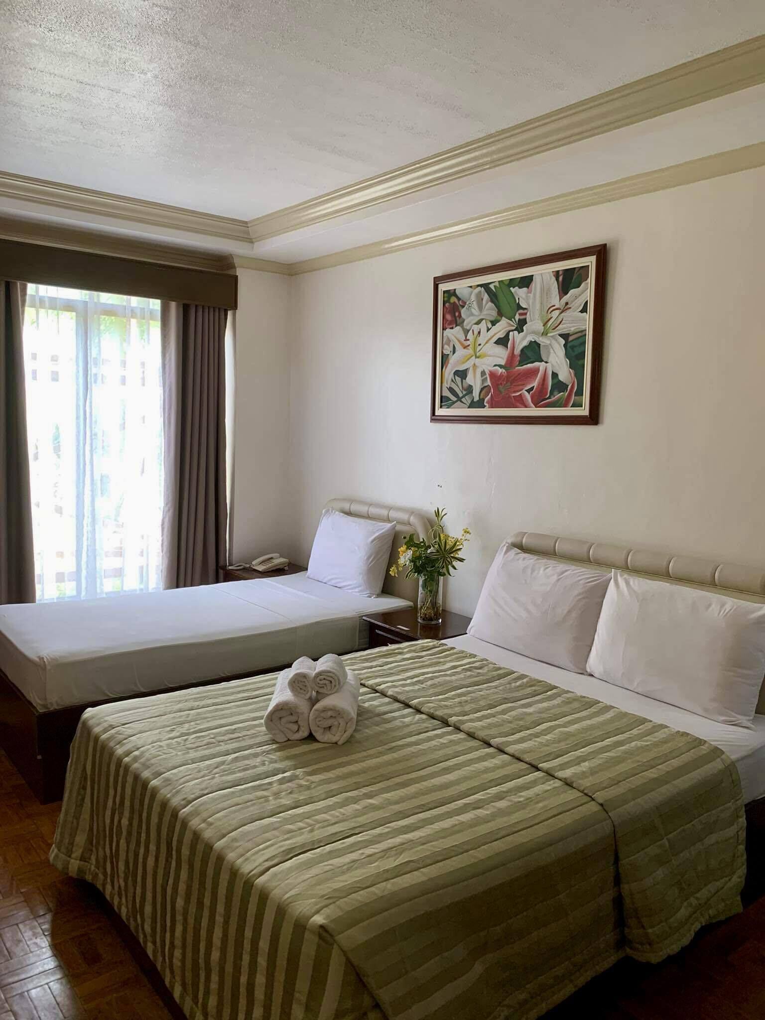 A hotel room with two beds, a large window with curtains, a painting on the wall, and a vase with flowers on the nightstand.