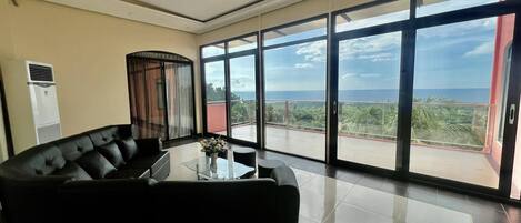 A modern living room with a black leather sofa, a glass coffee table, and a view of the ocean through large sliding glass doors.