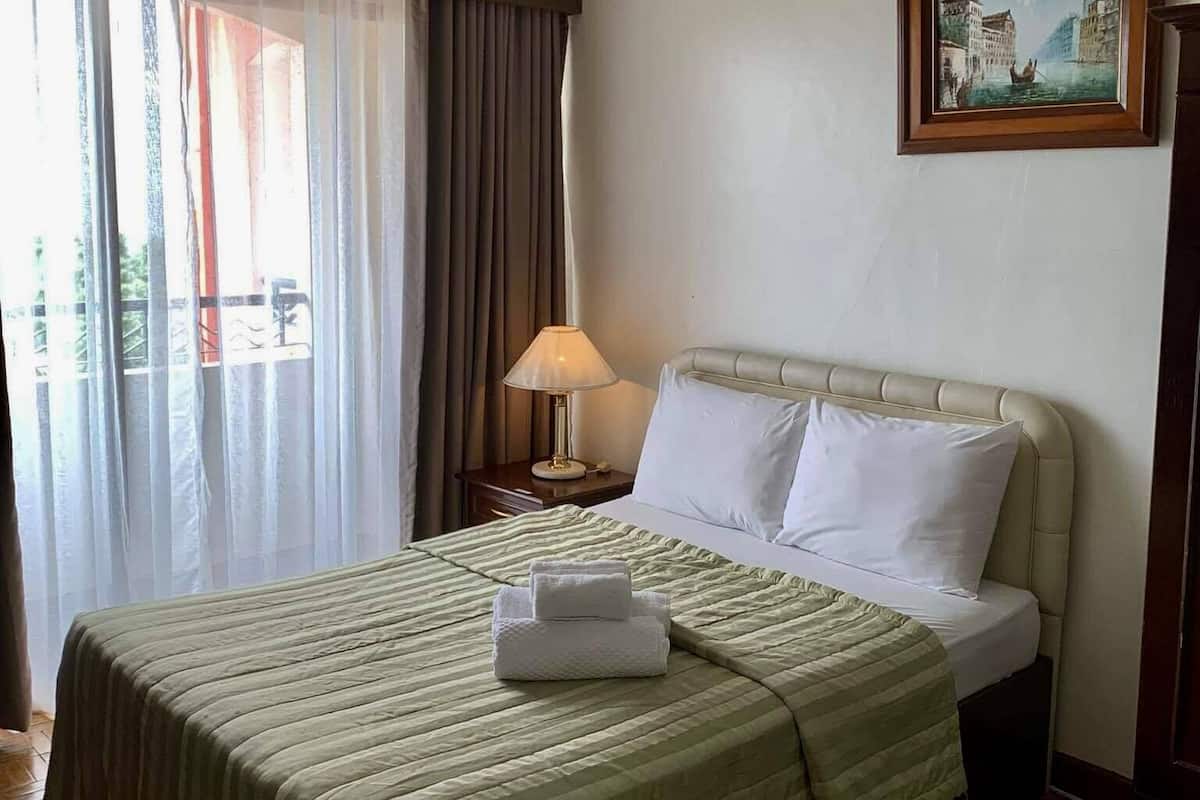 A hotel room with a bed, a nightstand, a lamp, a painting, and a view of the outdoors.