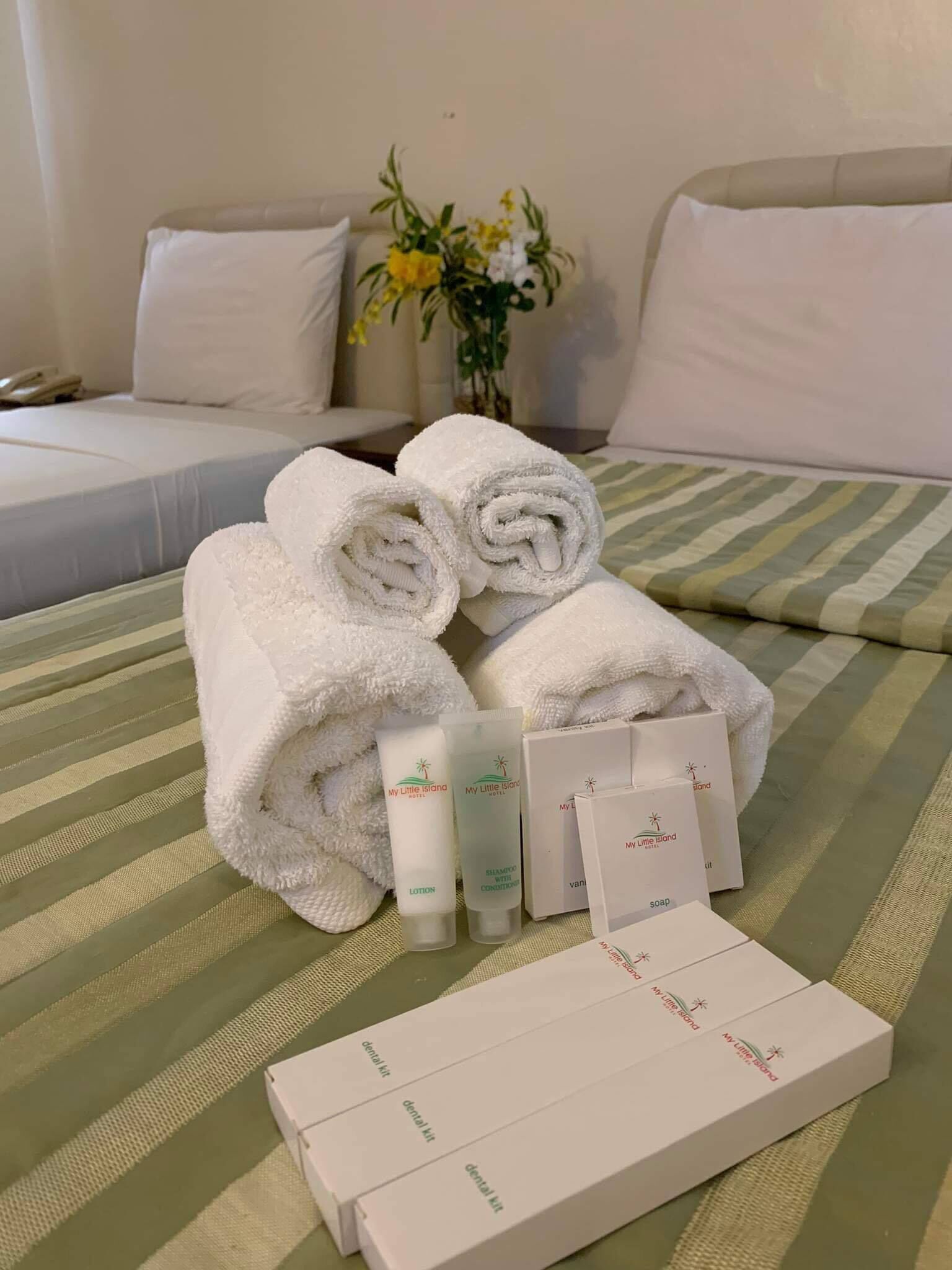 Una cama bien hecha con toallas blancas enrolladas, artículos de aseo como loción y jabón, y un kit de higiene dental sobre un edredón a rayas.