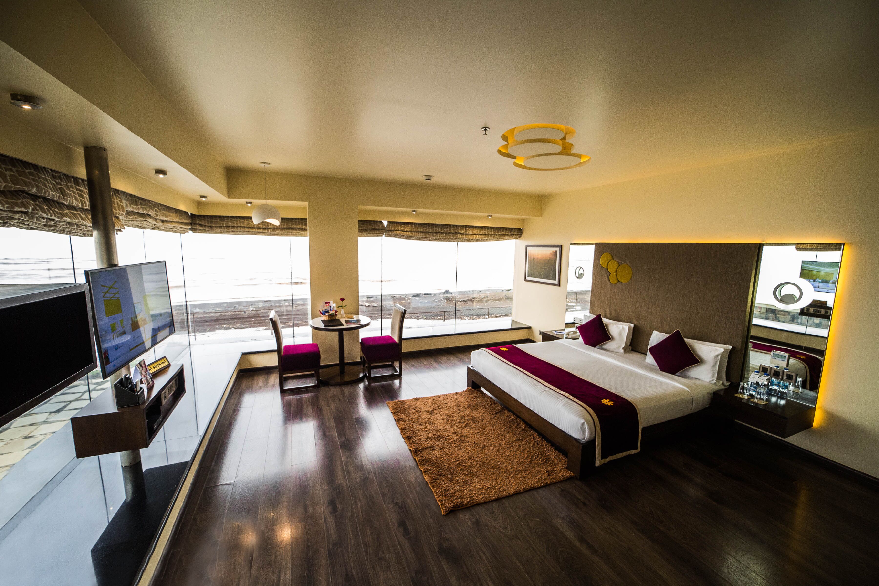 Quarto de hotel moderno com uma cama grande, televisão, uma mesinha com cadeiras e vista para o oceano.