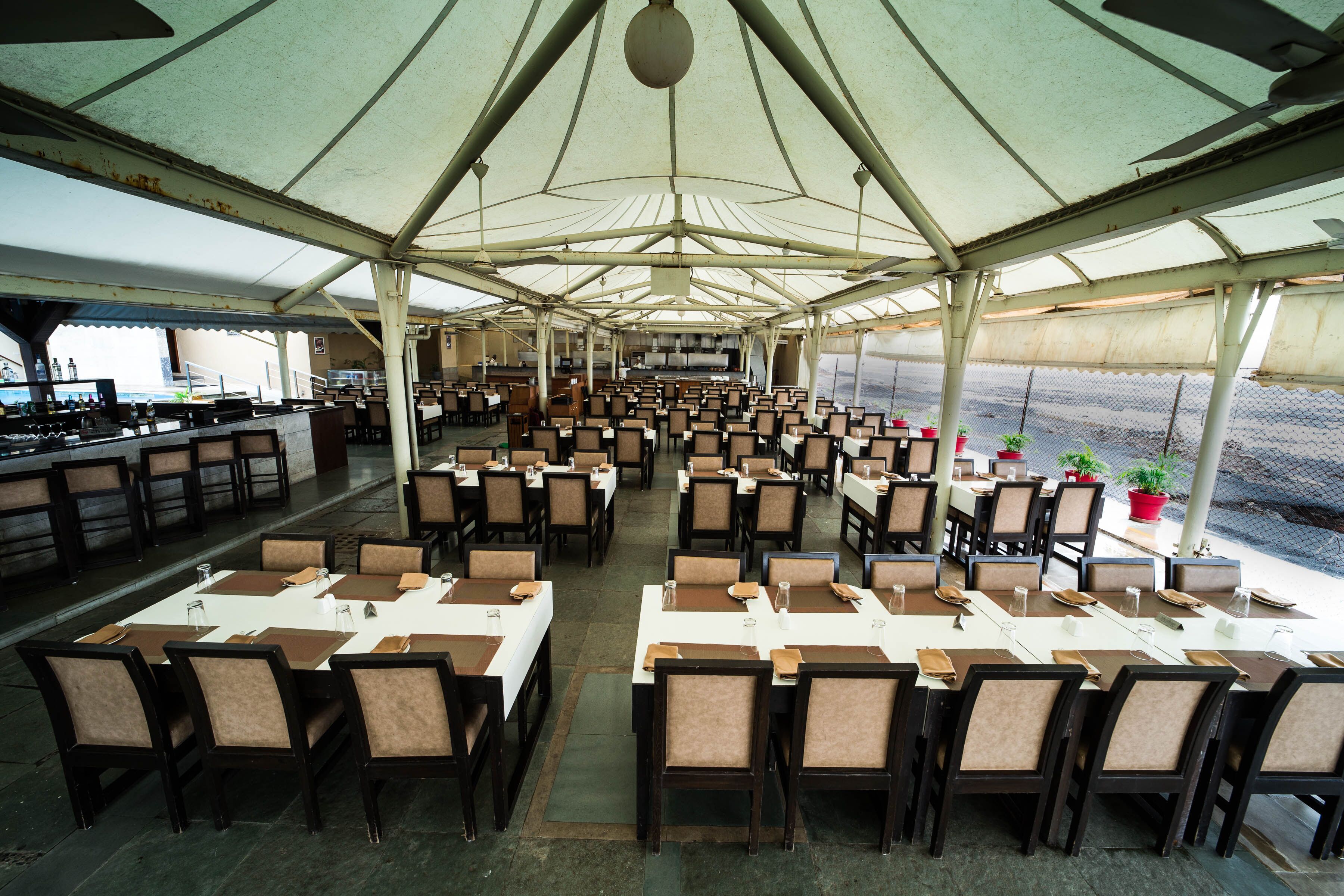 Un grand espace de restauration couvert, avec de nombreuses tables et chaises, un bar situé à gauche et des plantes en pot à droite.