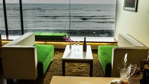 Una habitación con vista al océano, con asientos tapizados en verde, una mesita con una botella y copas, y un cuadro en la pared.