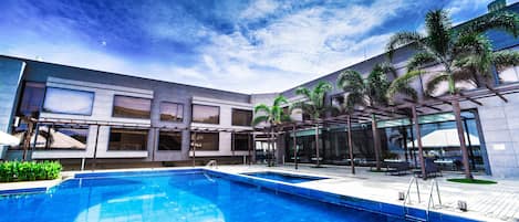 A modern hotel with a swimming pool, outdoor seating, and palm trees.