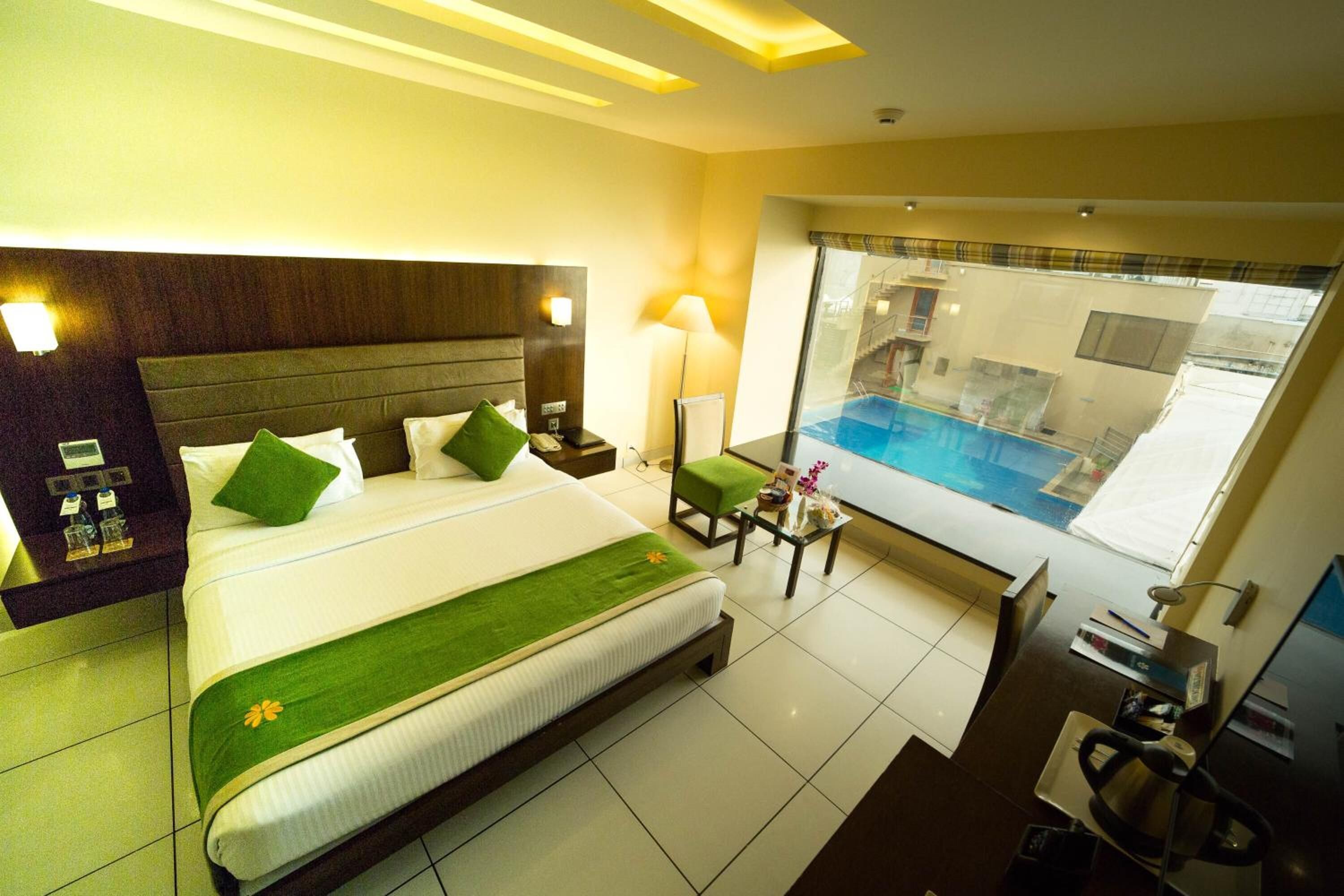 Ein Hotelzimmer mit einem großen Bett, einem kleinen Tisch mit Lampe, einem Sessel und Blick auf einen Pool.