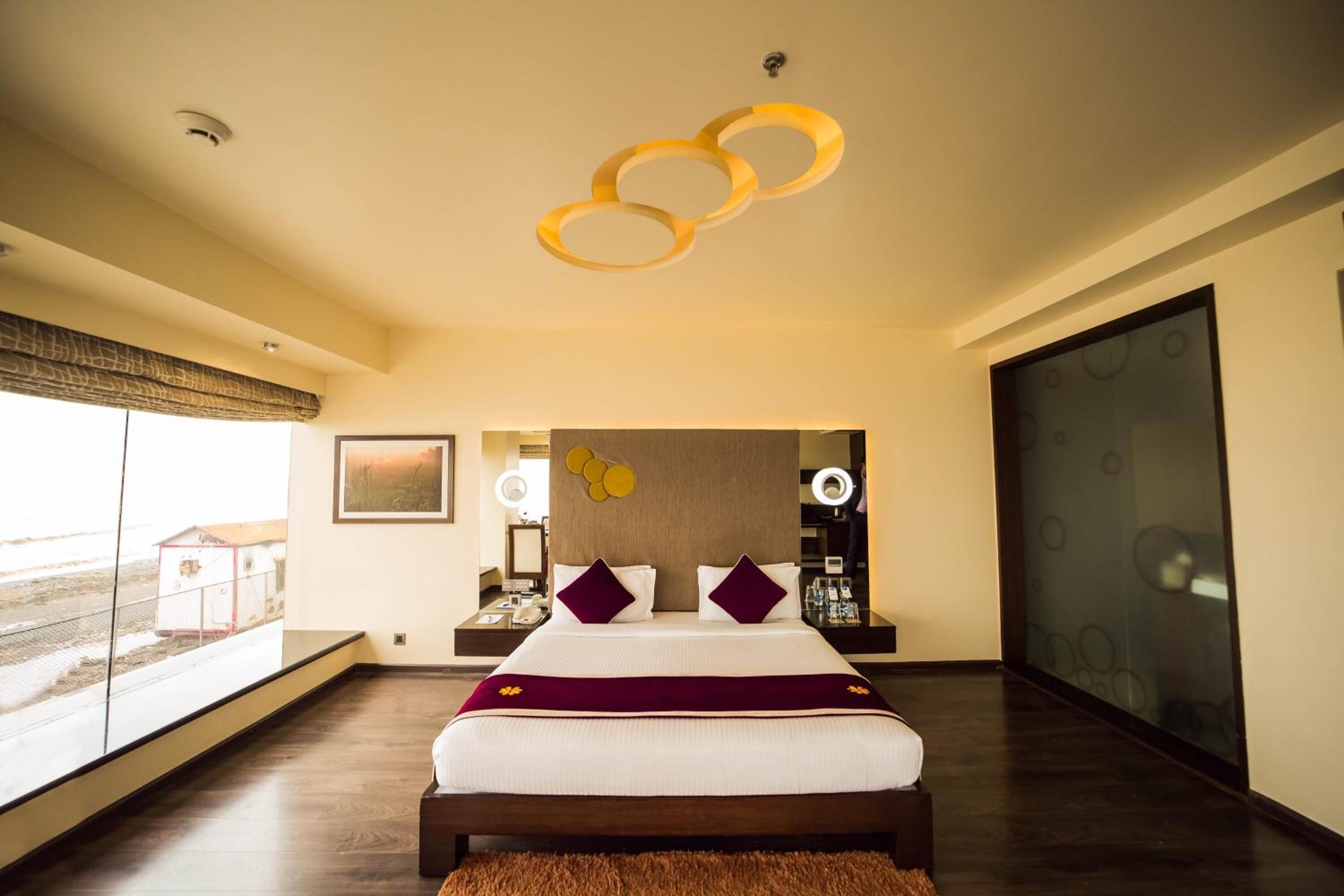 Ein modernes Hotelzimmer mit einem großen Bett, Blick durch ein Fenster und einer einzigartigen, runden Leuchte darüber.