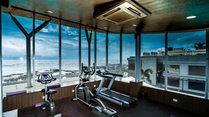 Un gimnasio con cinta caminadora y máquina elíptica, amplias ventanas con vista a la playa y una vista de un edificio al otro lado de la calle.