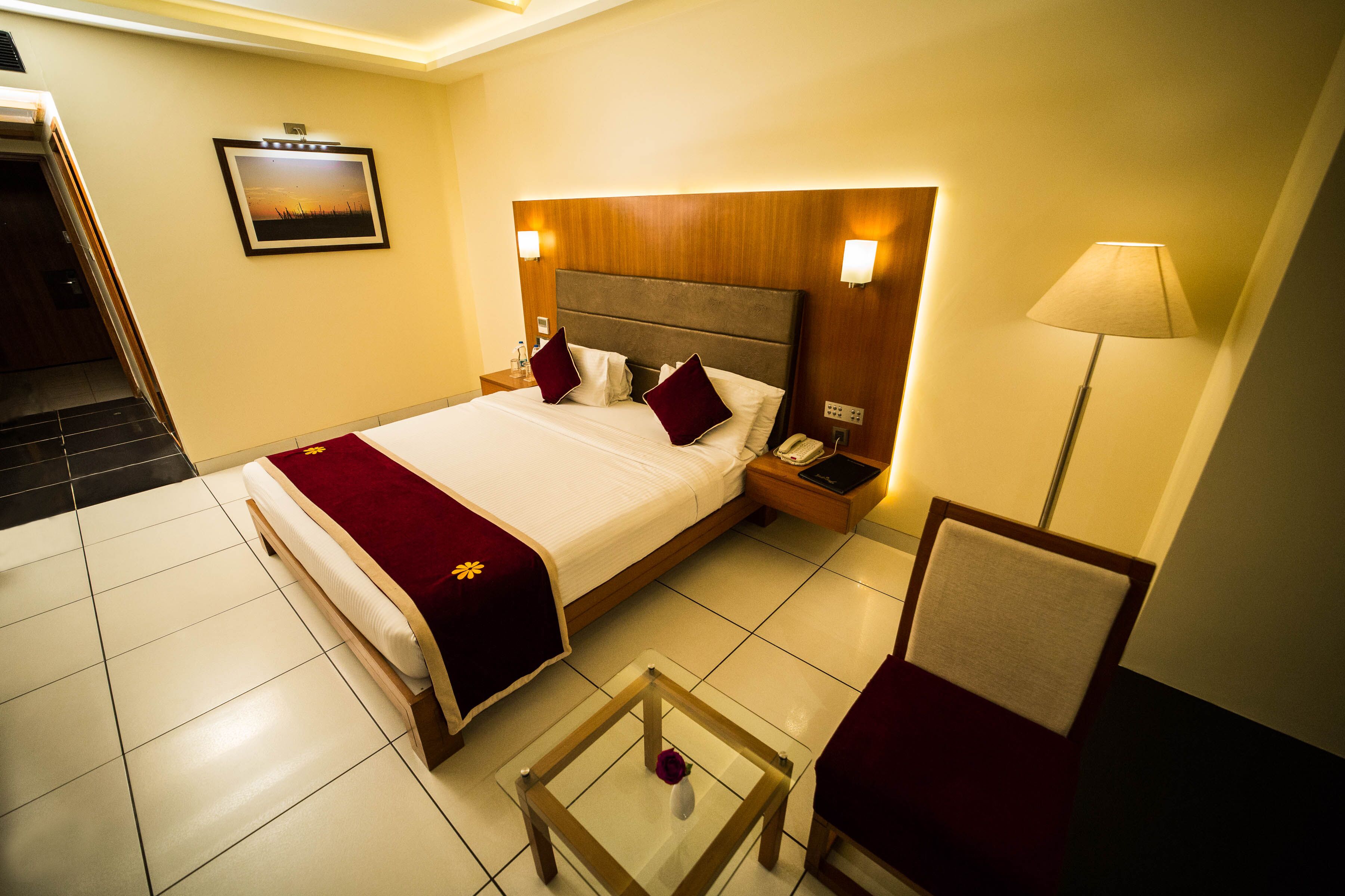 A hotel room with a large bed, a nightstand, a chair, a glass table with a vase, and a wall-mounted lamp.