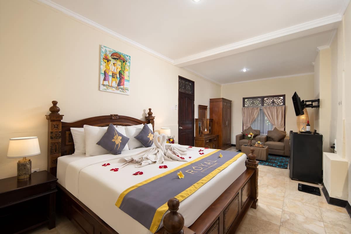 A hotel room with a large bed, a wooden headboard, a nightstand, a desk, a chair, a sofa, a refrigerator, and a window with curtains.