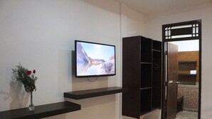 A room with a mounted TV on a wall, a dark wooden cabinet, and a small vase with flowers on a shelf. - Bali Senia Hotel (Denpasar)