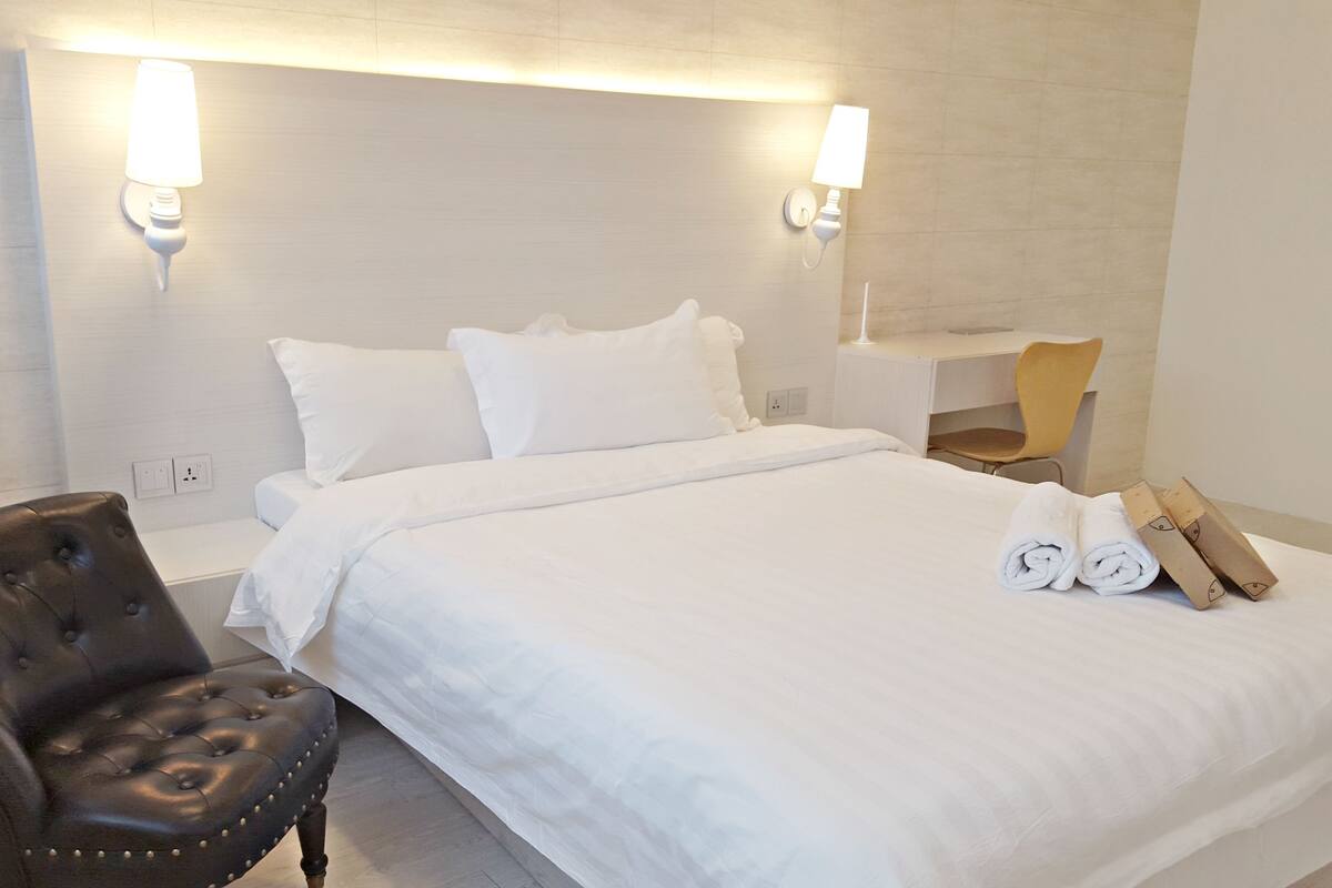 A hotel room with a neatly made bed, a leather armchair, a desk with a chair, and a rolled towel on the bed.