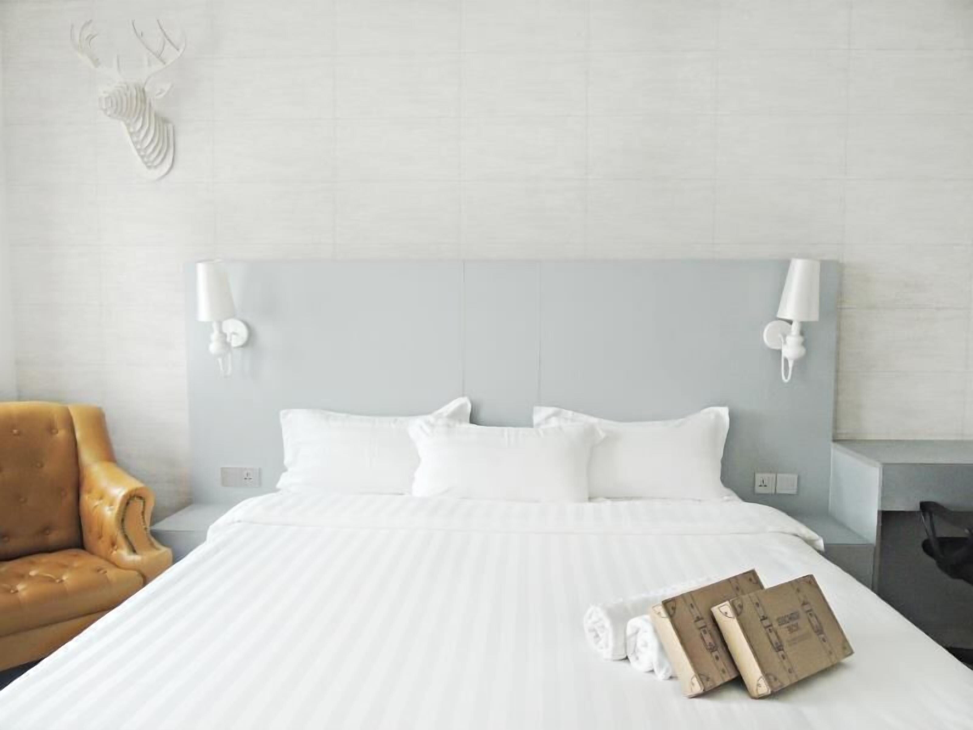 A hotel room with a bed, white pillows, a headboard, two wall-mounted lamps, a brown leather armchair, and rolled towels on the bed.