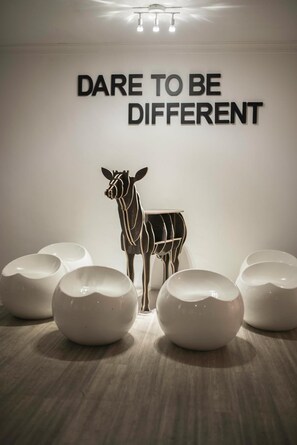 A wall with the text "DARE TO BE DIFFERENT" and a zebra sculpture. There are five white, round, modern stools in front. - Creator Hotel (Kulai)