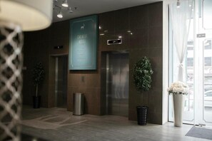 A hotel lobby with a reception desk, two elevators, a large poster, and potted plants.