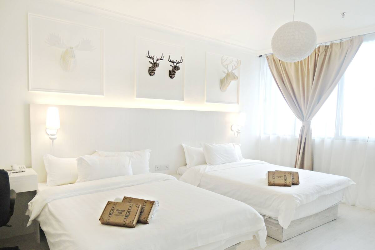 A hotel room with two beds, white bedding, a desk with a chair, and a shower box on each bed.