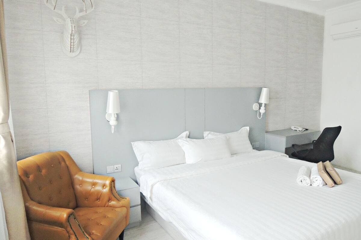 A hotel room with a large bed, a leather armchair, a nightstand with a lamp, and a wall-mounted deer head decoration.