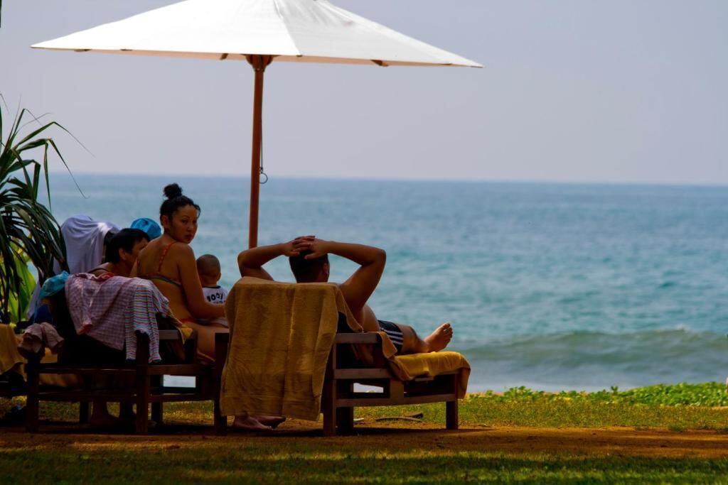 A group of people relaxing on wooden lounge chairs under a large umbrella by the ocean.