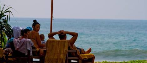 A group of people relaxing on wooden lounge chairs under a large umbrella by the ocean.