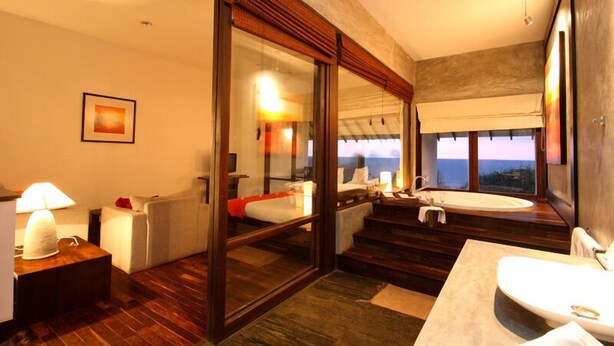 A modern hotel room with a large window, a bathtub, a sofa, and a desk with a lamp. - Temple Tree Resort & Spa (Induruwa)