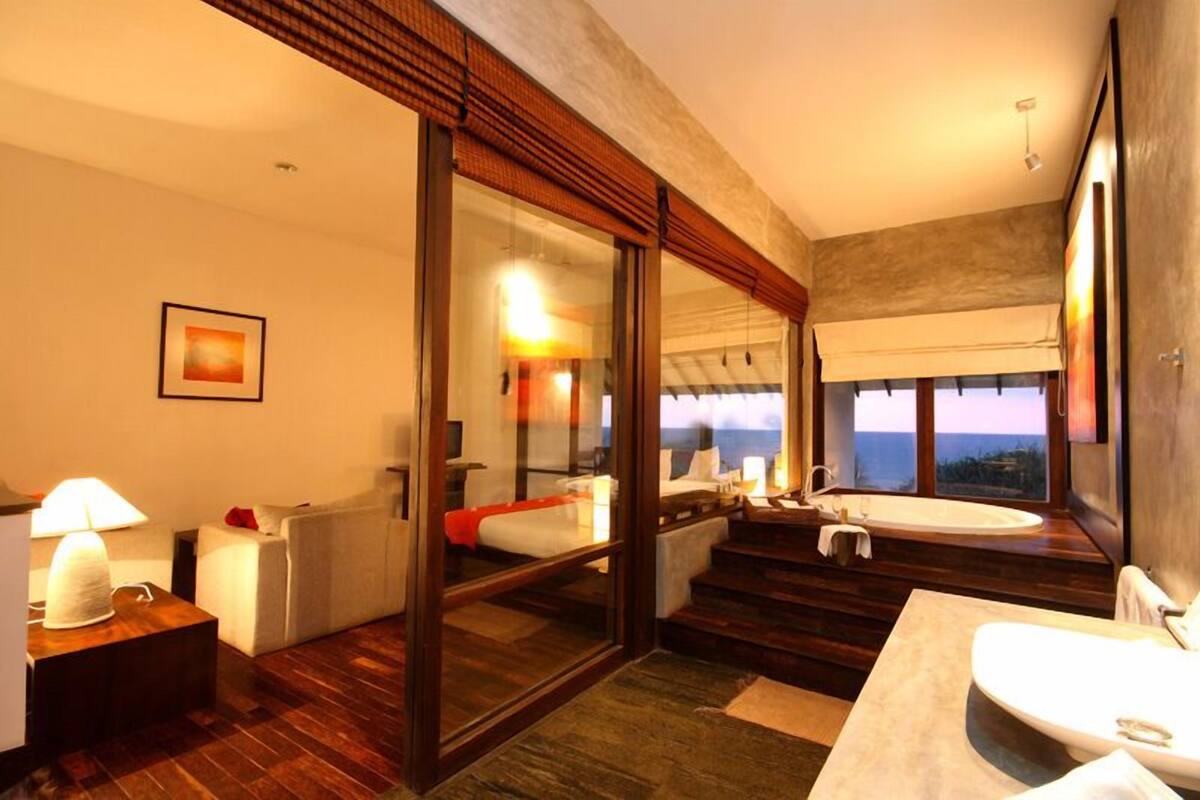 A modern hotel room with a large window, a bathtub, a sofa, and a desk with a lamp.
