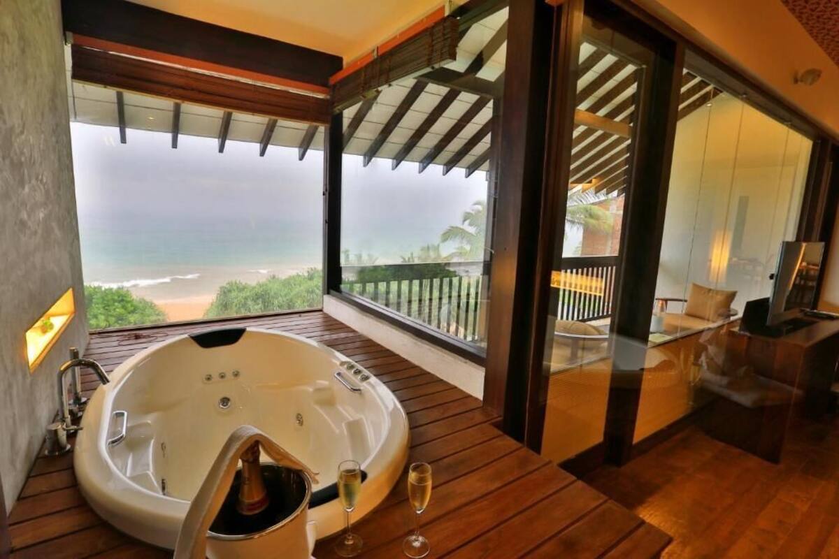 A room with a large bathtub, a balcony with a view of the beach, and a seating area with a TV.