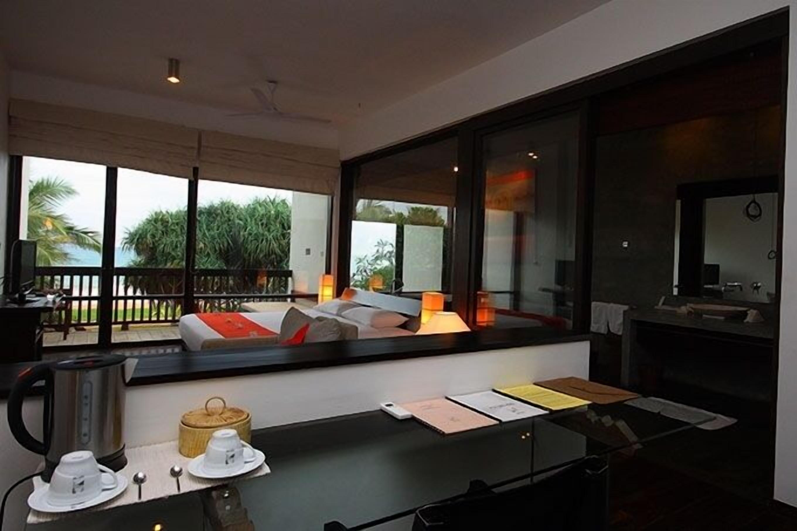 A modern hotel room with a large window offering a view of palm trees and a balcony. There is a dining area with a table, chairs, and a kettle.